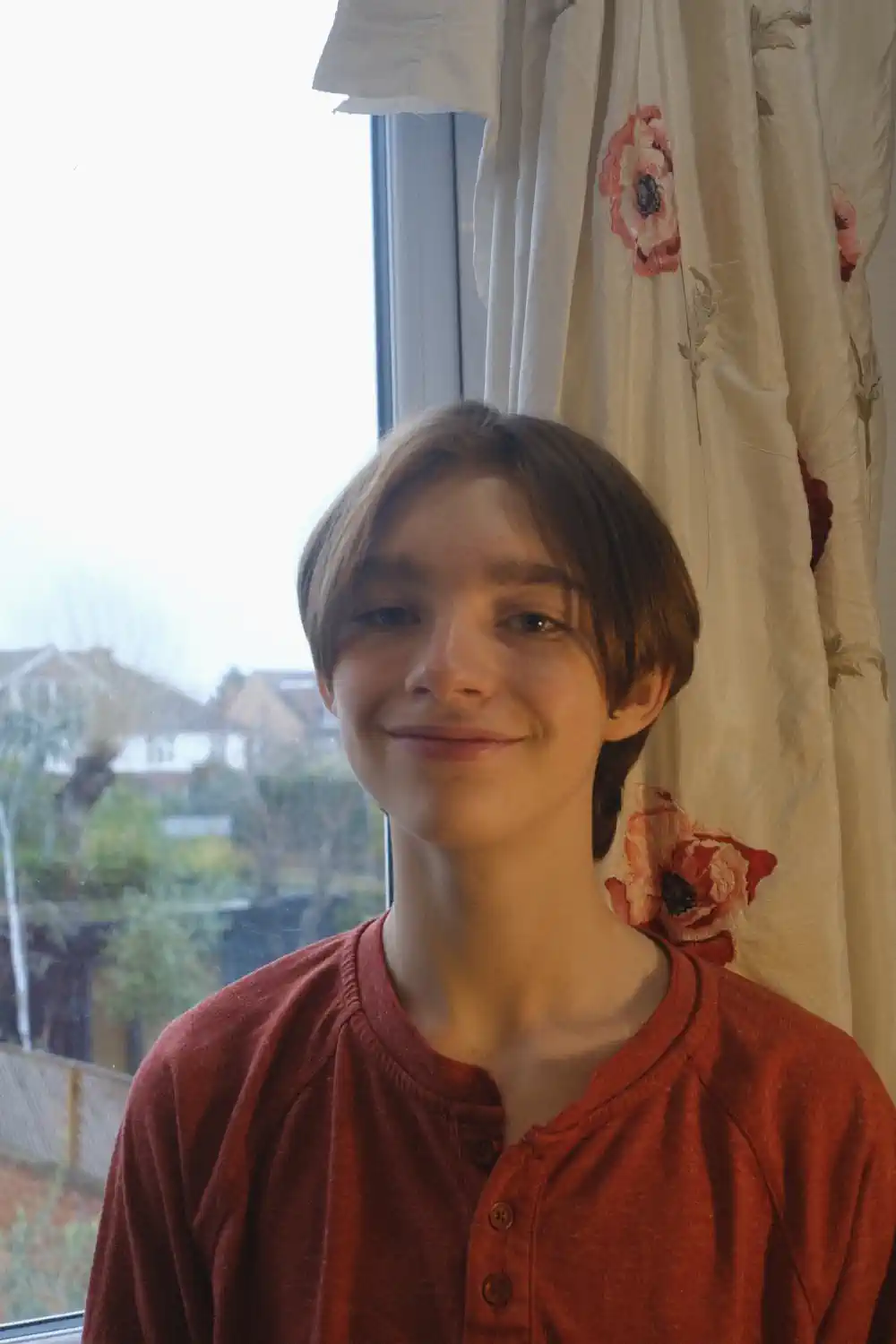 A picture of Gabriel KS, brown hair, blue eyes, white skin, smiling. Wearing a red shirt in front of a window 